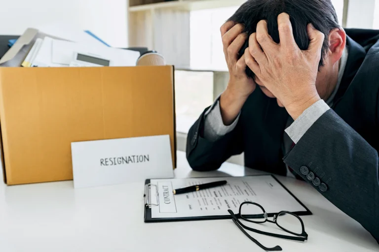 Un homme qui se tient la tête dans ses mains à cîté d'un carton d'affaires et une lettre de résignation